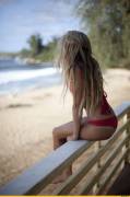 Blonde perched on railing in red cheeky bikini watching the ocean waves