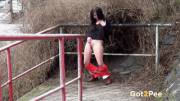 Dark haired girl stands next to a railing and pulls down her trousers to piss